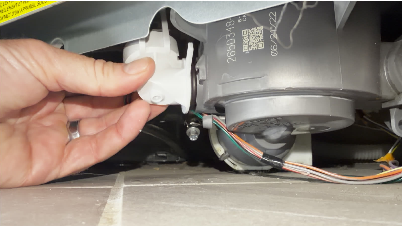 Reaching under the dishwasher with the kick plate removed, gripping the white pressure sensor at the sump