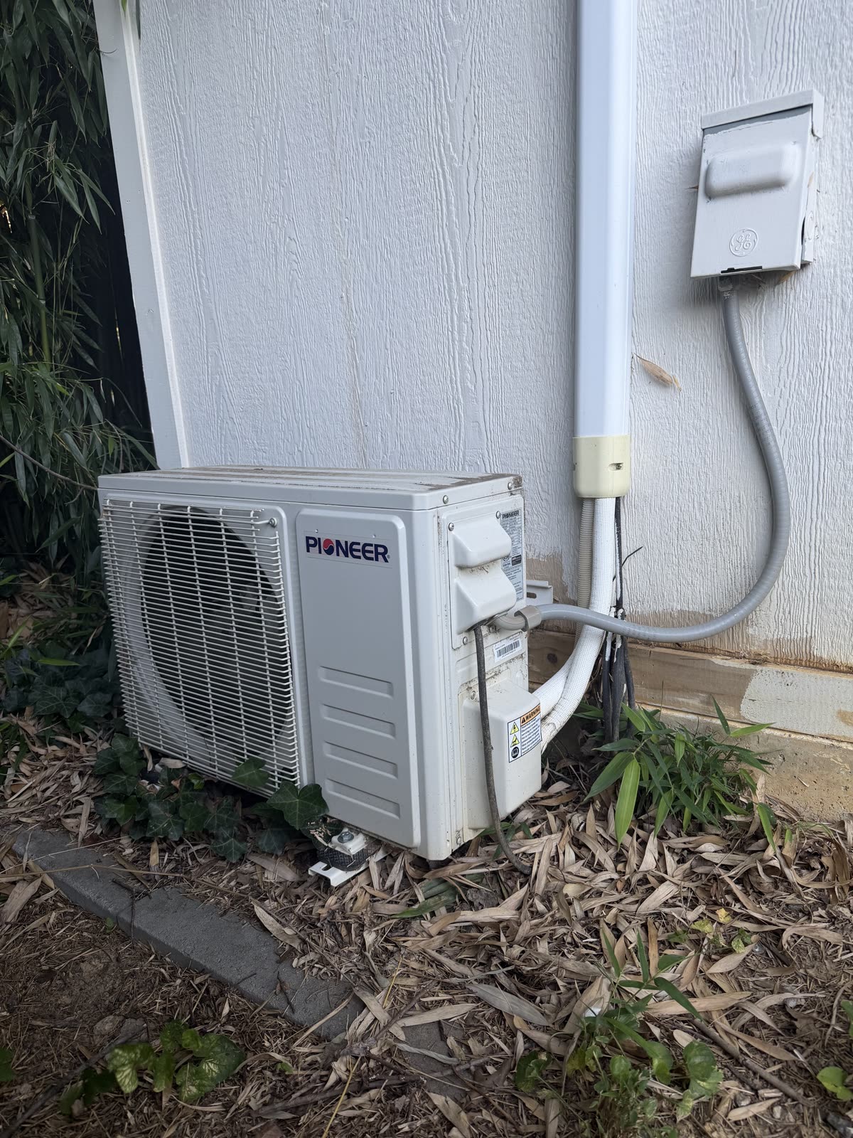 Mini split outdoor unit beside the house with a conduit whip running to a wall-mounted disconnect box